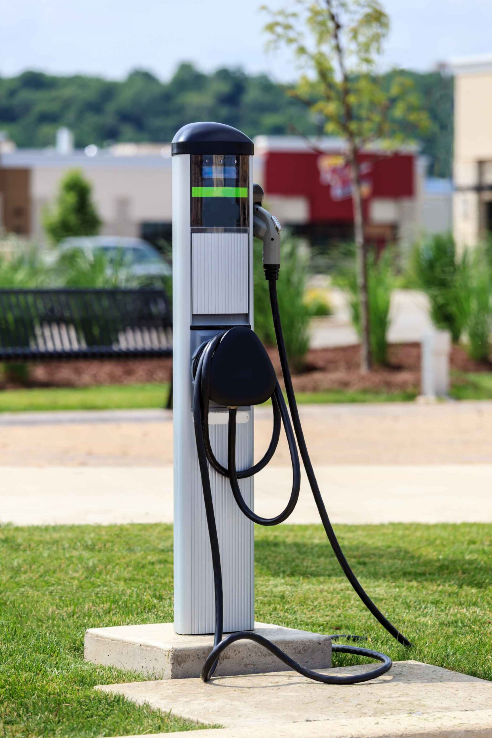 Outdoor electric vehicle charging station installed on a concrete base with dual charging cables, set in a landscaped urban area.